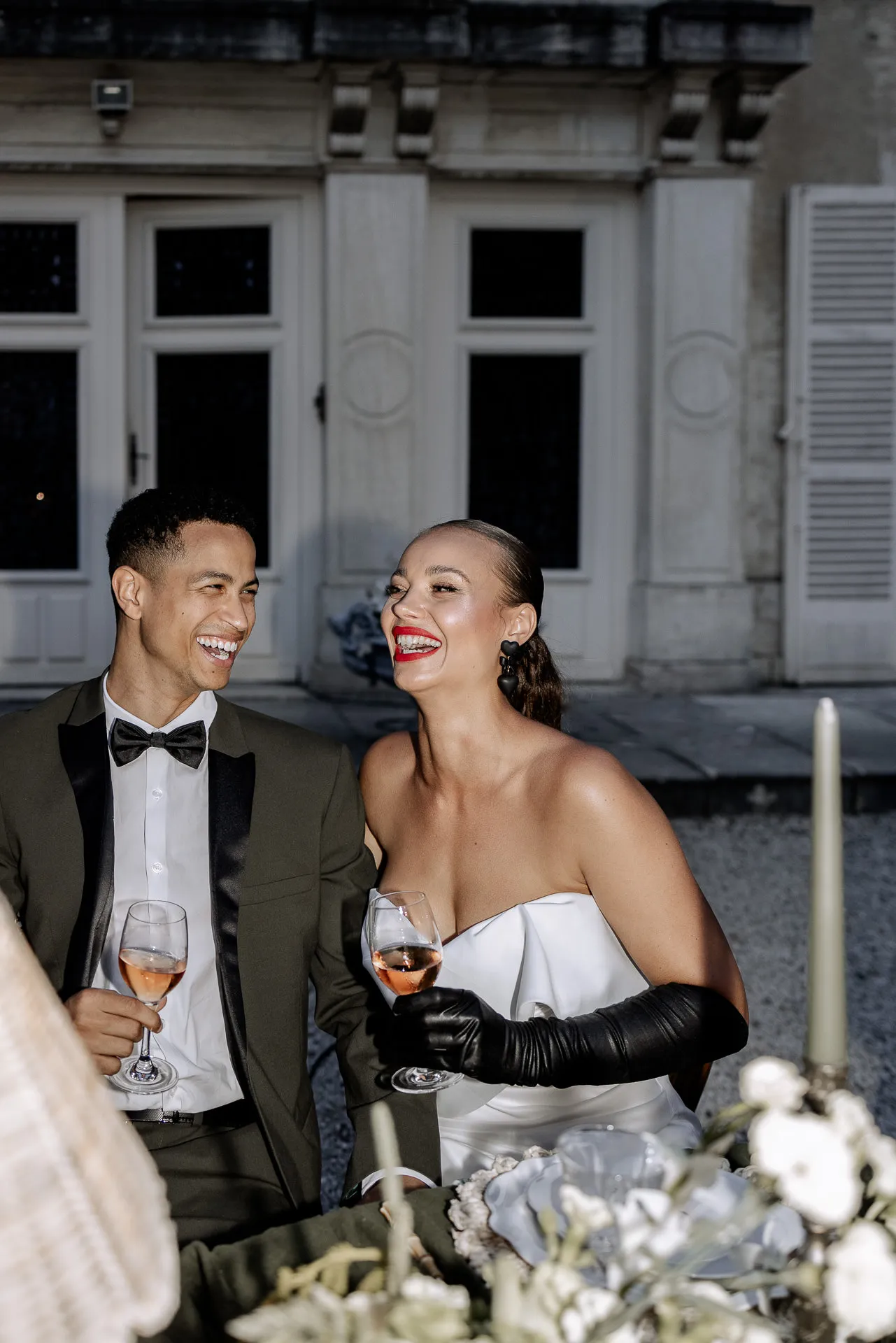Lachendes Brautpaar mit Weingläsern bei Hochzeitsempfang vor dem Chateau de Varennes in Frankreich